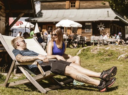 Entdecken Sie im Sommerurlaub die Tiroler Bergseen. Mann mit Tätowierung und Frau entspannen auf Liegestuhl vor rustikalem Gebäude im Sonnenschein