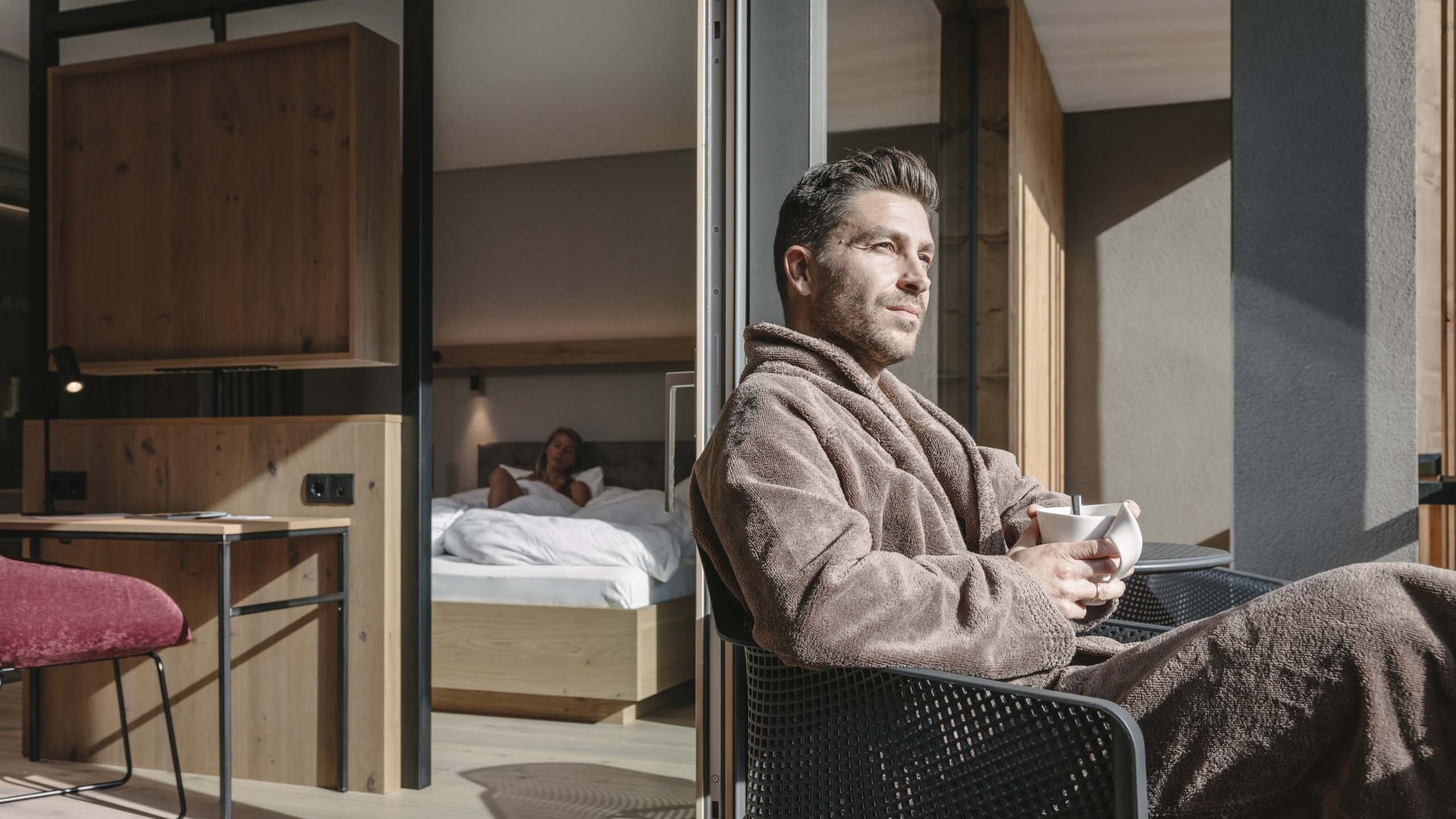 Feiern Sie Ihre Feste in Tirol Mann im Bademantel trinkt Kaffee auf Balkon, Frau liegt im Bett im Hintergrund