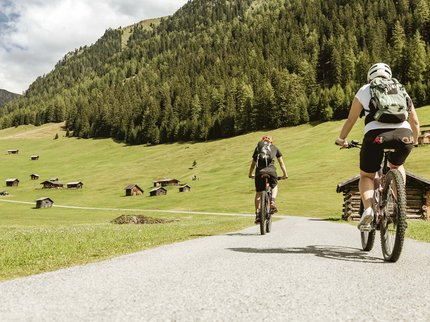 Ihr Radurlaub in Österreich/Tirol Zwei Radfahrer fahren auf Landstraße durch bergige Almlandschaft mit Holzhütten