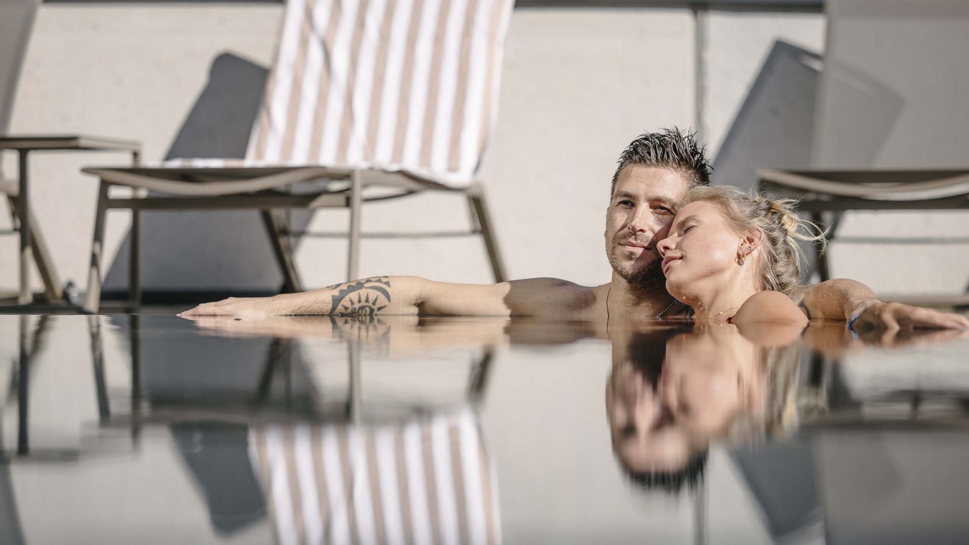 Wellnesshotel in Tirol gesucht? Paar relaxt zusammen am Pool mit Liegestühlen und Handtuch im Hintergrund