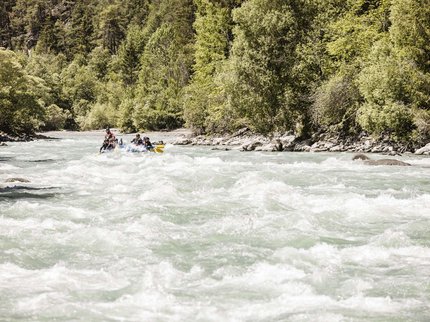 Entdecken Sie im Sommerurlaub die Tiroler Bergseen. Menschen beim Wildwasser-Rafting auf einem schnell fließenden Fluss umgeben von Bäumen
