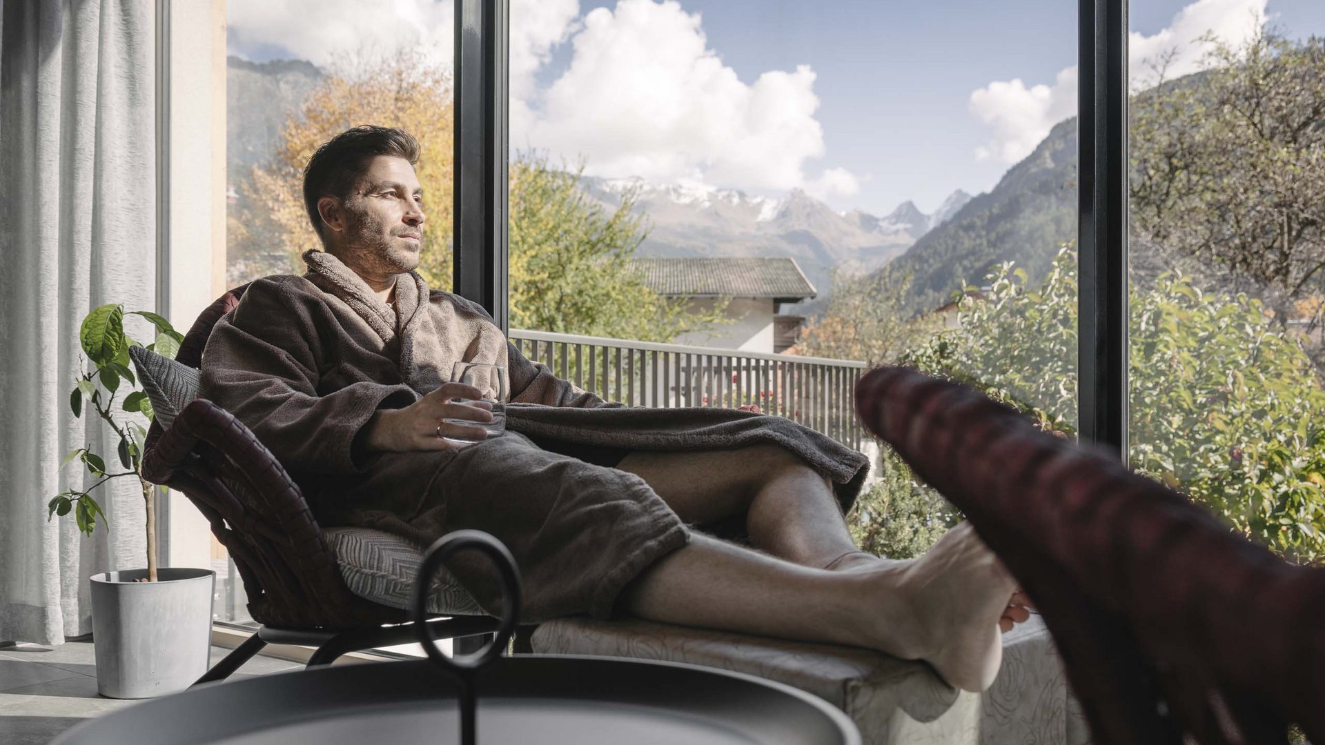 Hotel in Prutz gesucht? Mann in Bademantel entspannt sich mit Getränk und Bergblick durch großes Fenster