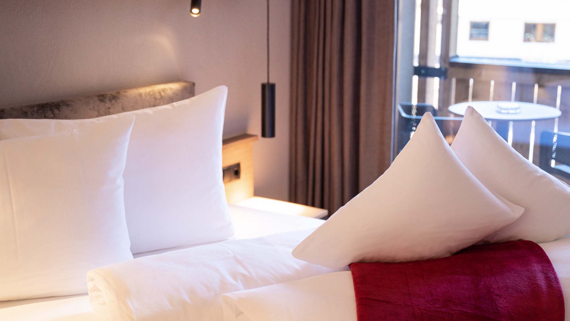 Ihr Hotel im Tiroler Oberland Modernes Hotelzimmer mit weißer Bettwäsche und roten Akzenten am Fenster
