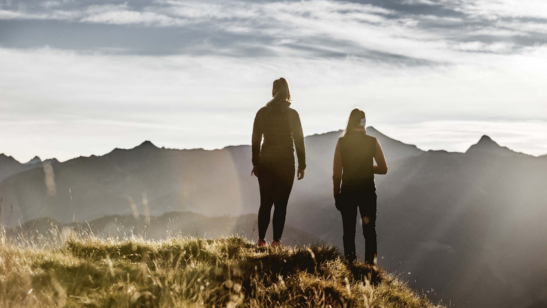 Ihr Urlaub im Sporthotel in Tirol Zwei Personen stehen auf einem grasbewachsenen Hügel mit Bergpanorama im Hintergrund