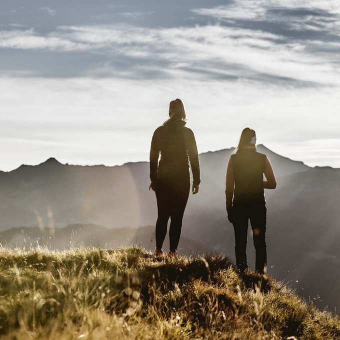 Entdecken Sie im Sommerurlaub die Tiroler Bergseen. Zwei Personen stehen auf einem grasbewachsenen Hügel mit Bergpanorama im Hintergrund