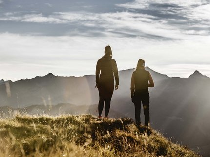 Entdecken Sie im Sommerurlaub die Tiroler Bergseen. Zwei Personen stehen auf einem grasbewachsenen Hügel mit Bergpanorama im Hintergrund