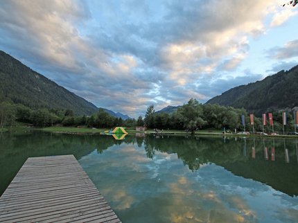 Entdecken Sie im Sommerurlaub die Tiroler Bergseen. Holzsteg am ruhigen See mit Bergen und Wolken am Himmel