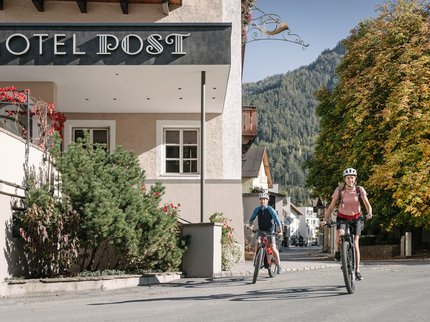 Entdecken Sie im Sommerurlaub die Tiroler Bergseen. Zwei Radfahrer vor Hotel Post in einer Stadt mit Bergen und Herbstbäumen