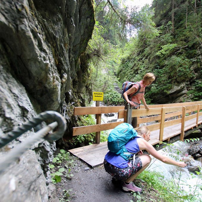 Entdecken Sie im Sommerurlaub die Tiroler Bergseen. Zwei Wanderinnen auf einem Waldpfad an einem Fluss mit einer Holzbrücke