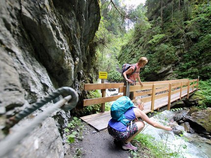 Entdecken Sie im Sommerurlaub die Tiroler Bergseen. Zwei Wanderinnen auf einem Waldpfad an einem Fluss mit einer Holzbrücke