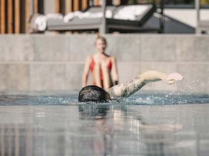 Wellnesshotel in Tirol gesucht? Person mit Tattoo schwimmt im Pool, andere Person sitzt im Hintergrund