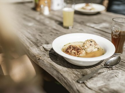 Entdecken Sie im Sommerurlaub die Tiroler Bergseen. Knödel in Brühe auf rustikalem Holztisch mit Getränk und Löffel