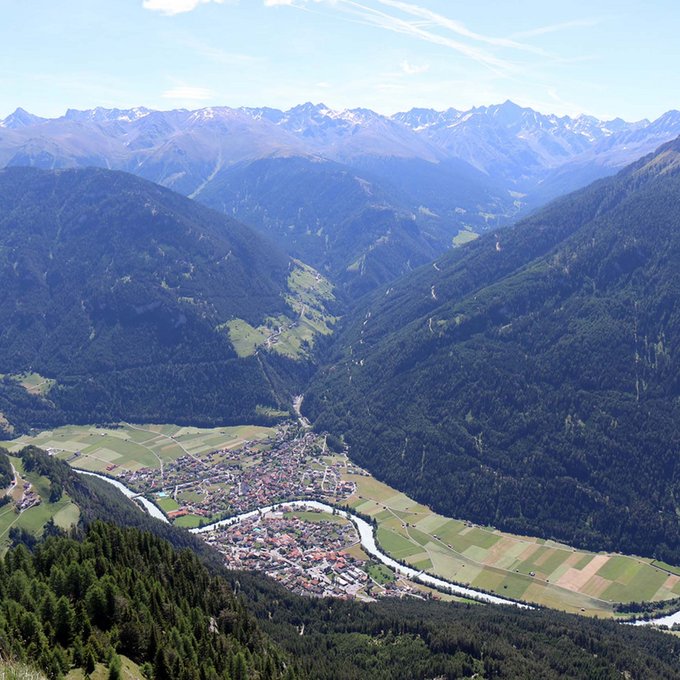Motorradtouren in Tirol Blick auf ein Tal mit Fluss und Dorf umgeben von bewaldeten Bergen und schneebedeckten Gipfeln