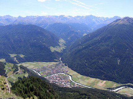 Entdecken Sie im Sommerurlaub die Tiroler Bergseen. Blick auf ein Tal mit Fluss und Dorf umgeben von bewaldeten Bergen und schneebedeckten Gipfeln