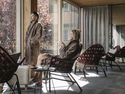 Ihr Urlaub im Sporthotel in Tirol Mann steht und Frau sitzt in Bademänteln am großen Fenster in einer modernen Lounge