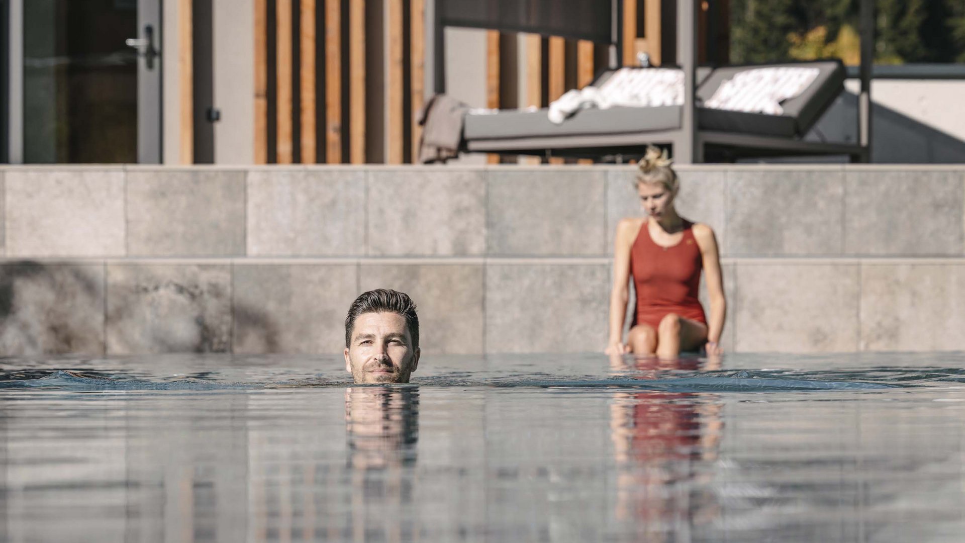 Outdoorpool Mann schwimmt im Pool, Frau sitzt am Beckenrand im roten Badeanzug
