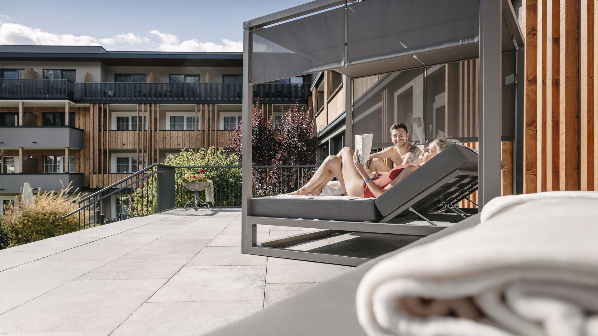 Wellnesshotel in Tirol gesucht? Paar entspannt auf Liege mit Sonnenschutz auf Terrasse eines modernen Gebäudes
