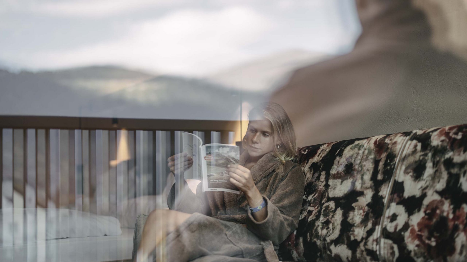 Ihr Hotel im Tiroler Oberland Frau liest auf einem Sofa mit Blumendruck, durch ein Fenster mit Reflexionen fotografiert