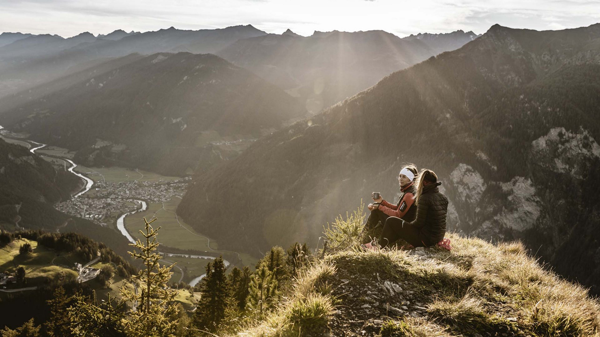 Ihr Wohlfühlort: das Hotel in Prutz im Tiroler Oberland ⛰️ Zwei Wanderer sitzen auf Berggipfel mit Tal- und Flussblick bei Sonnenstrahlen
