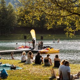 Ihr Wohlfühlort: das Hotel in Prutz im Tiroler Oberland ⛰️ Menschen sitzen am Seeufer und hören Live-Musik auf einem Steg im Sonnenschein